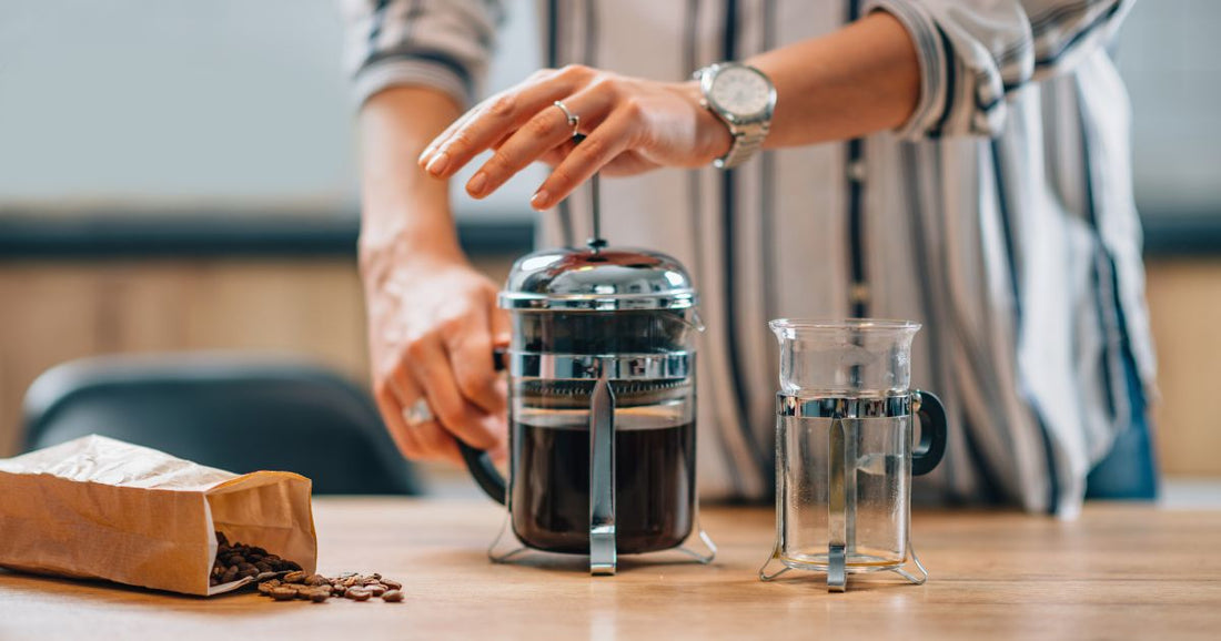 Person pressing a French press coffee maker on a table beside coffee beans and an empty class coffee cup